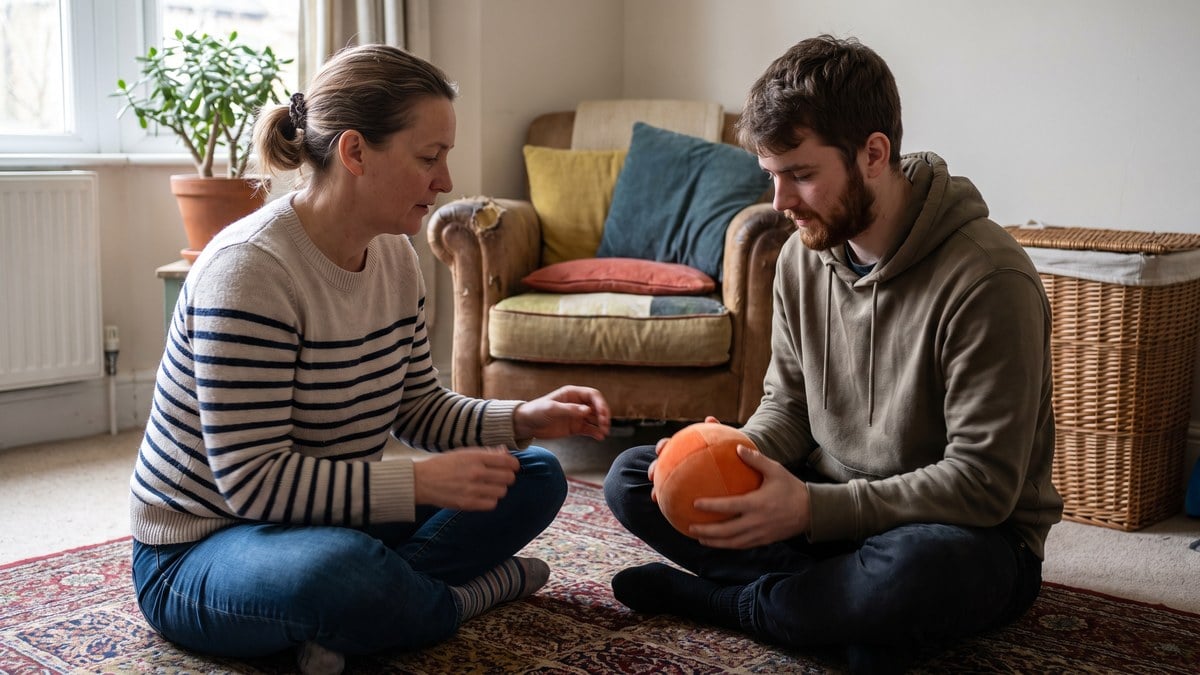 An occupational therapist sitting on a living room floor beside a young man, working through hand exercises with a soft ball